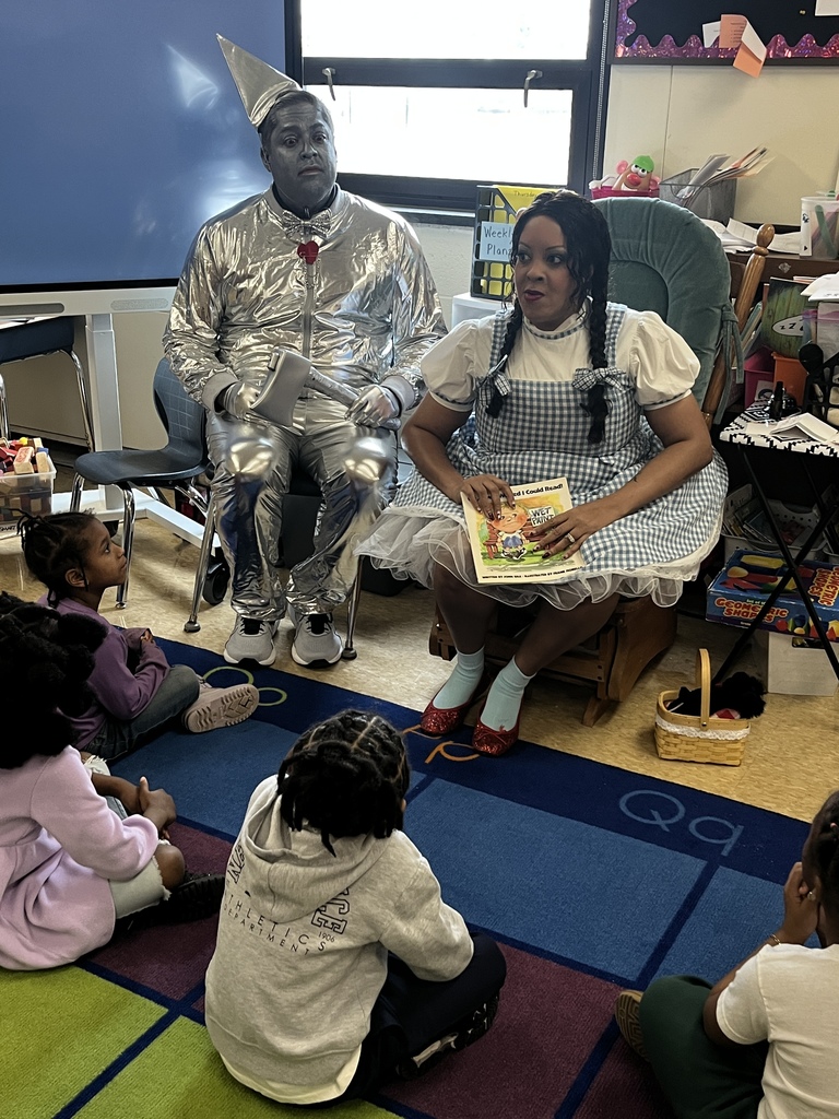 A guest reader dressed as Dorothy holds a book while speaking to students seated on the rug, with a Tin Man character sitting beside her.