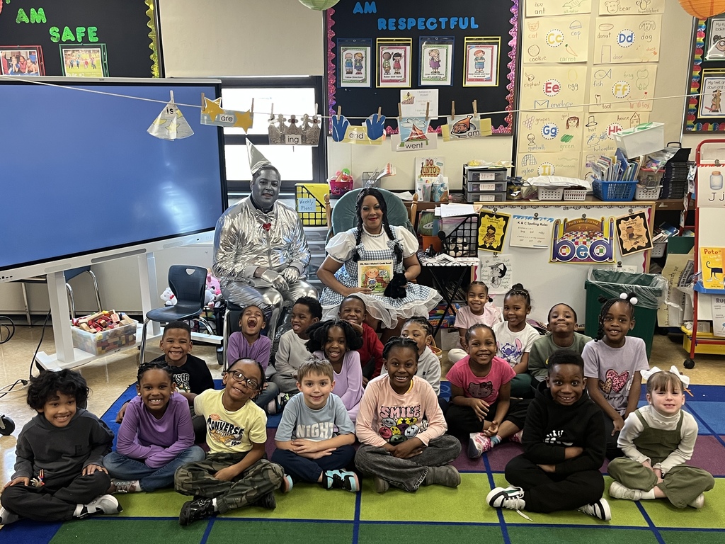 A class of smiling elementary students sits together on a classroom rug with two guest readers dressed as Dorothy and the Tin Man.