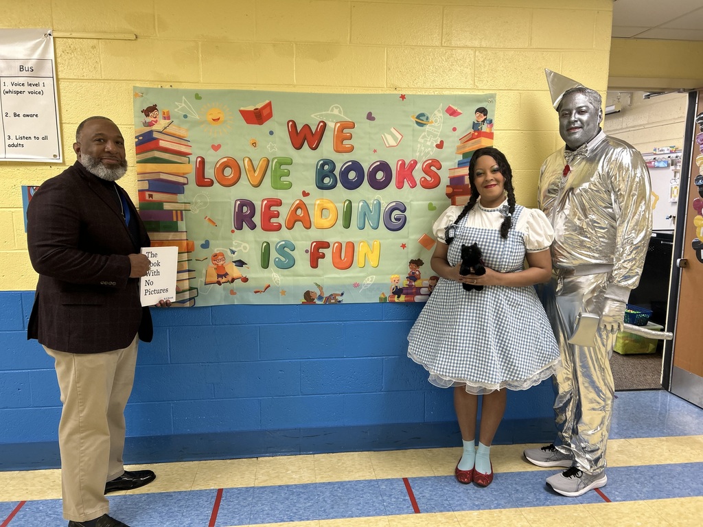 IMG_4069Three adults dressed as storybook characters stand in a school hallway beside a “We Love Books – Reading Is Fun” banner, promoting reading.