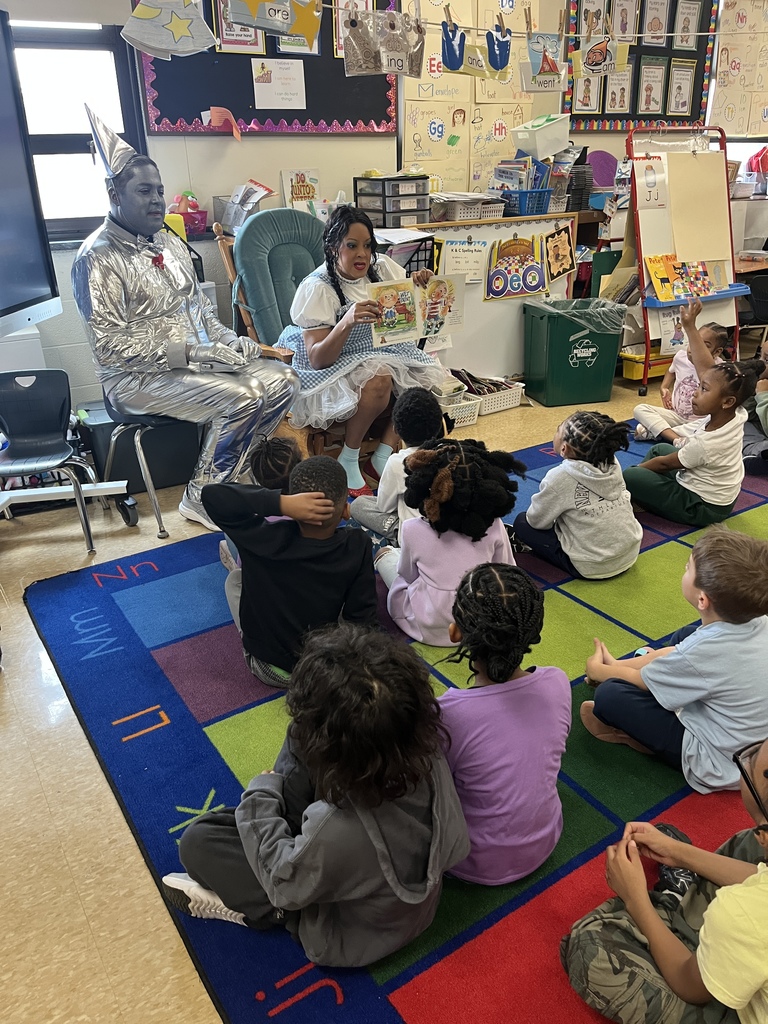 A guest reader dressed as Dorothy reads a picture book to elementary students sitting on a classroom rug while a Tin Man character sits nearby.