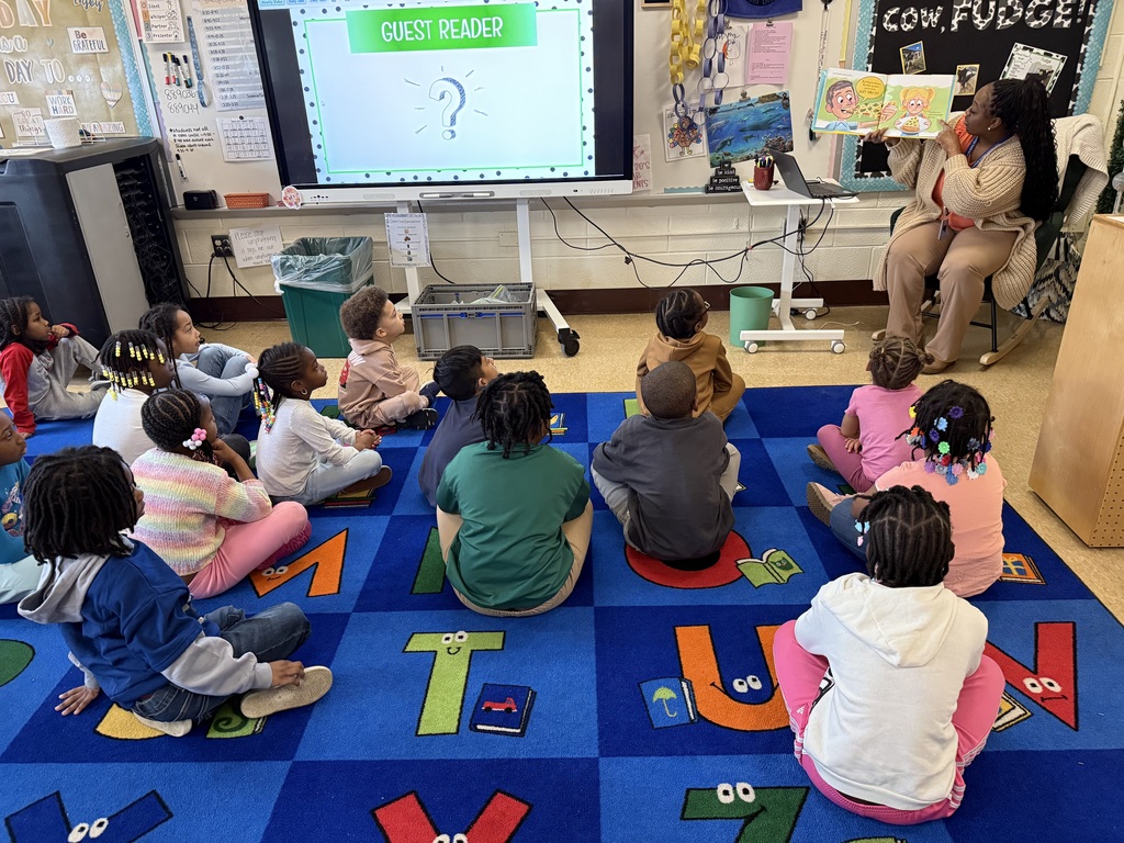 A teacher reads a picture book aloud to students seated on a classroom rug while a screen behind her displays “Guest Reader.”