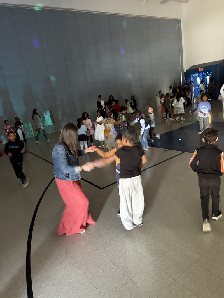 Children dancing and playing in a large indoor gymnasium with a woman in a pink skirt and denim jacket.