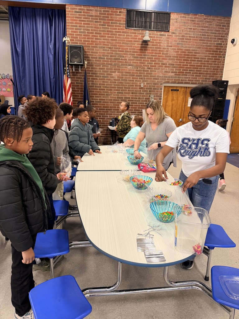 Students gather around a cafeteria table with bowls of candy, engaging in a group activity inside a school gymnasium.
