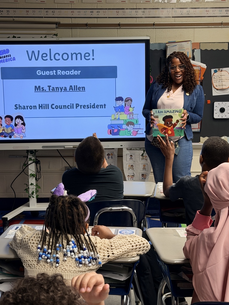 Guest reader Ms. Tanya Allen stands in front of a classroom holding a children's book while students attentively listen and raise hands.