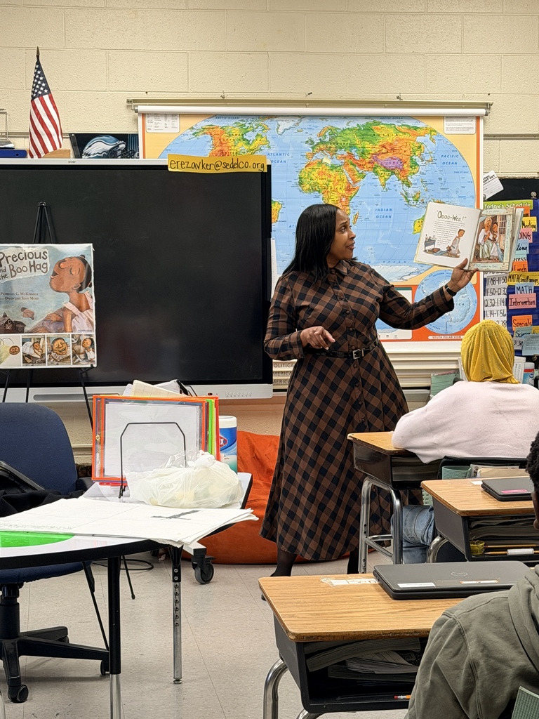 Teacher in a plaid dress reads a children's book aloud to attentive students in a colorful classroom with a world map and American flag.
