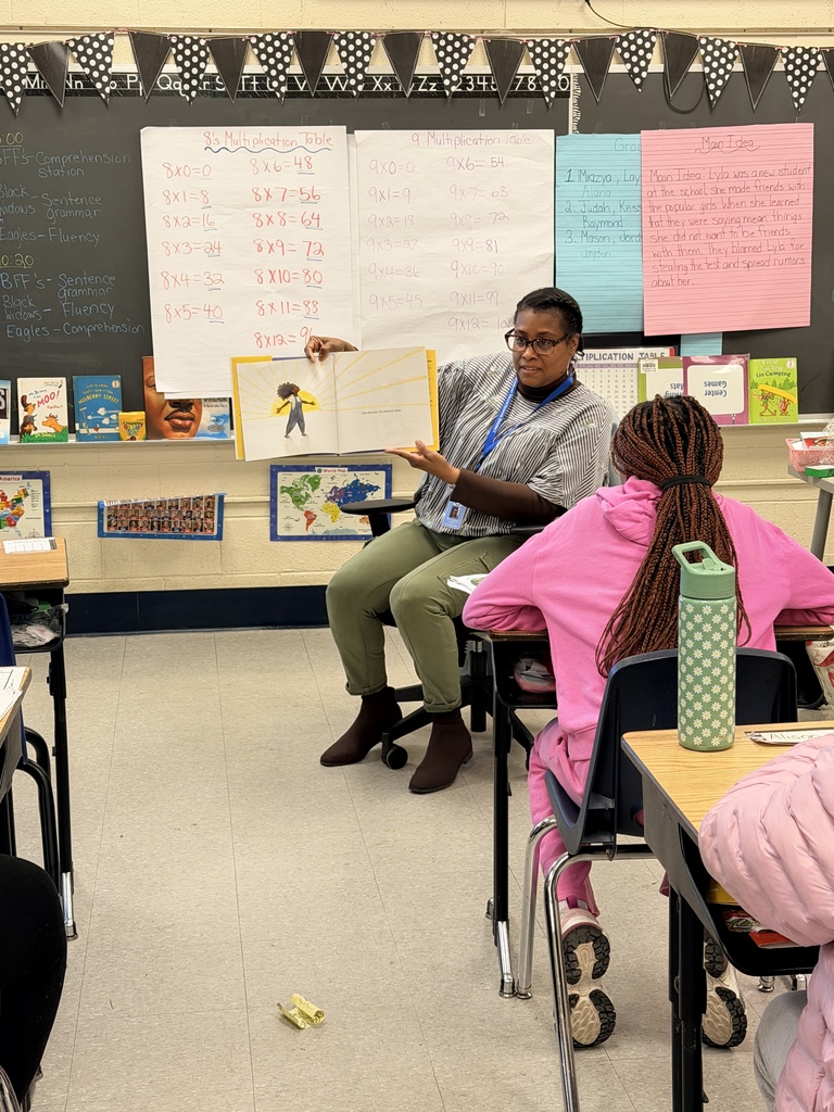 Teacher seated in front of a classroom reading a picture book aloud to attentive students at their desks.