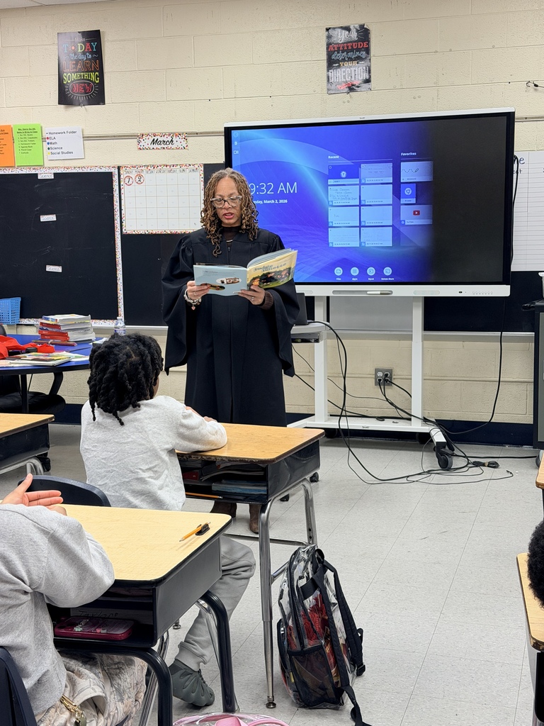 Teacher in a black robe reading a book aloud to attentive elementary students seated at desks in a classroom with a digital screen behind her.