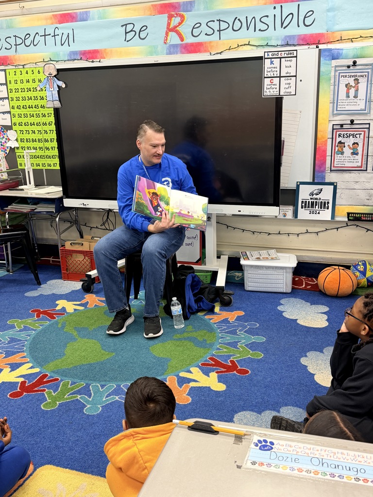 A person reads a colorful book aloud to children seated on a vibrant classroom rug with educational posters on the walls.