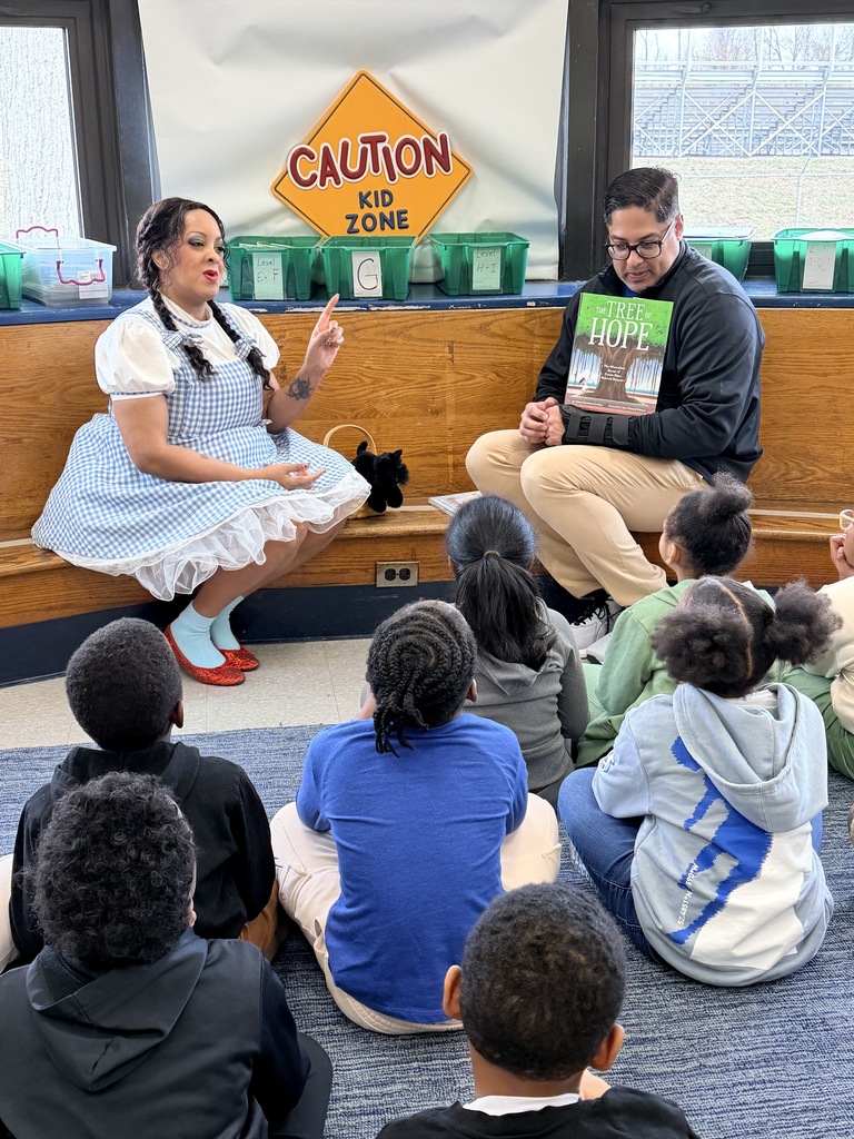 Woman dressed as Dorothy from The Wizard of Oz reading to children seated on the floor in a classroom with a 'Caution Kid Zone' sign behind her.