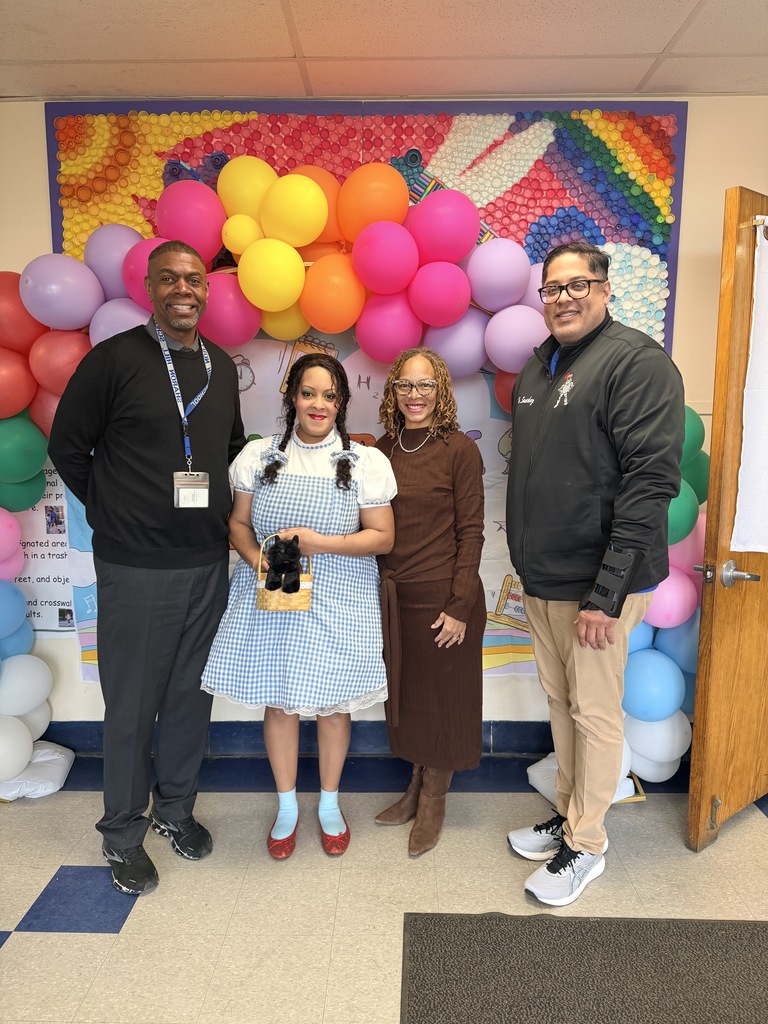 Four people stand in front of a colorful balloon arch with rainbow and heart decorations indoors.