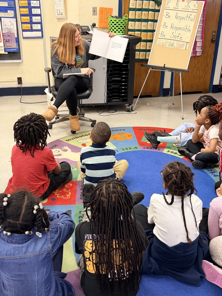 Teacher reading a book aloud to a group of attentive young children seated on a colorful classroom rug.