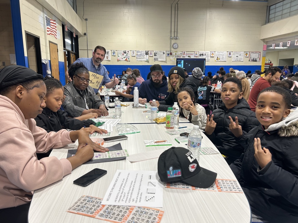 Students and families play bingo at cafeteria tables while a sign reads “50/50 for $1” during a school event.