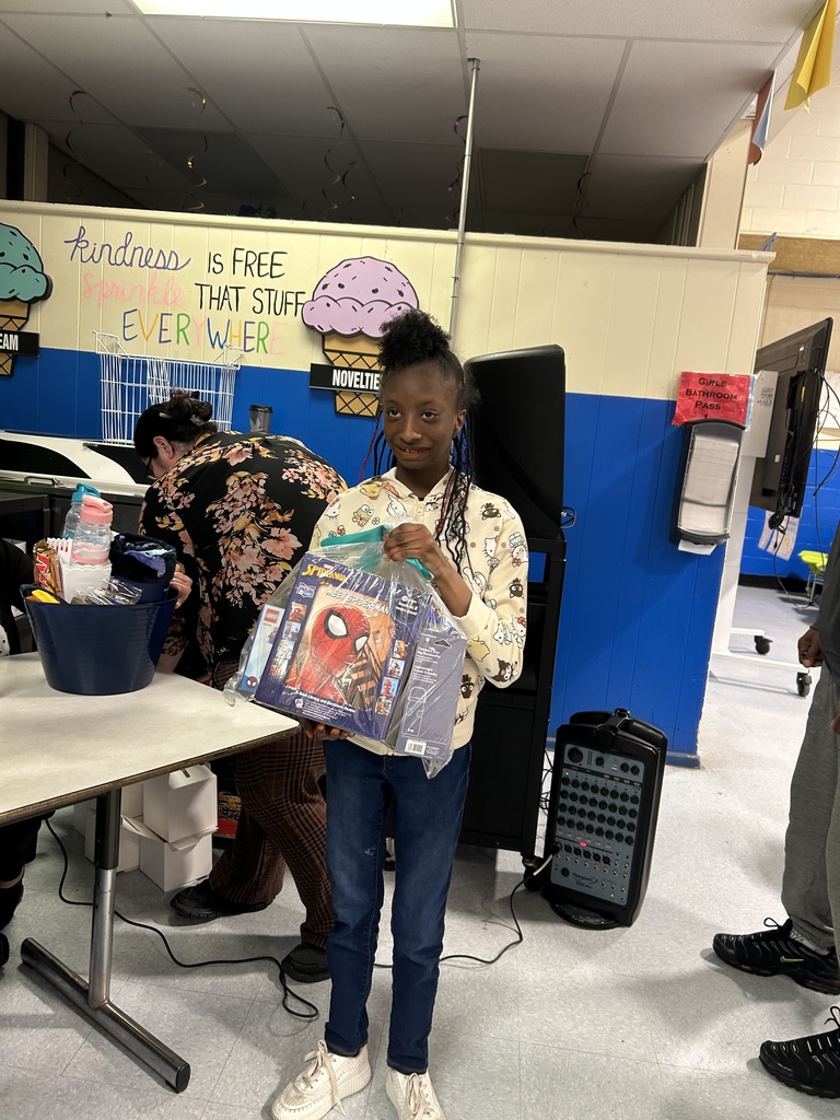 A student smiles while holding a bag with a Spider-Man-themed prize in a decorated school cafeteria.