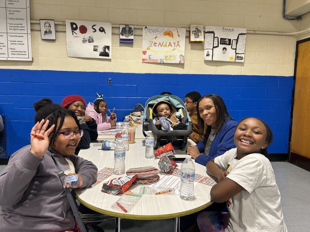 Families and students sit around a table with snacks and drinks, smiling and interacting during a school gathering.