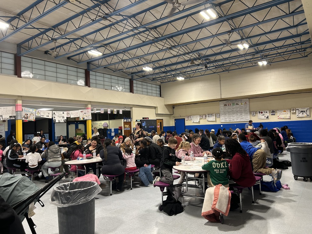 A large group of families and students fill a cafeteria, seated at tables and participating in a lively school event.