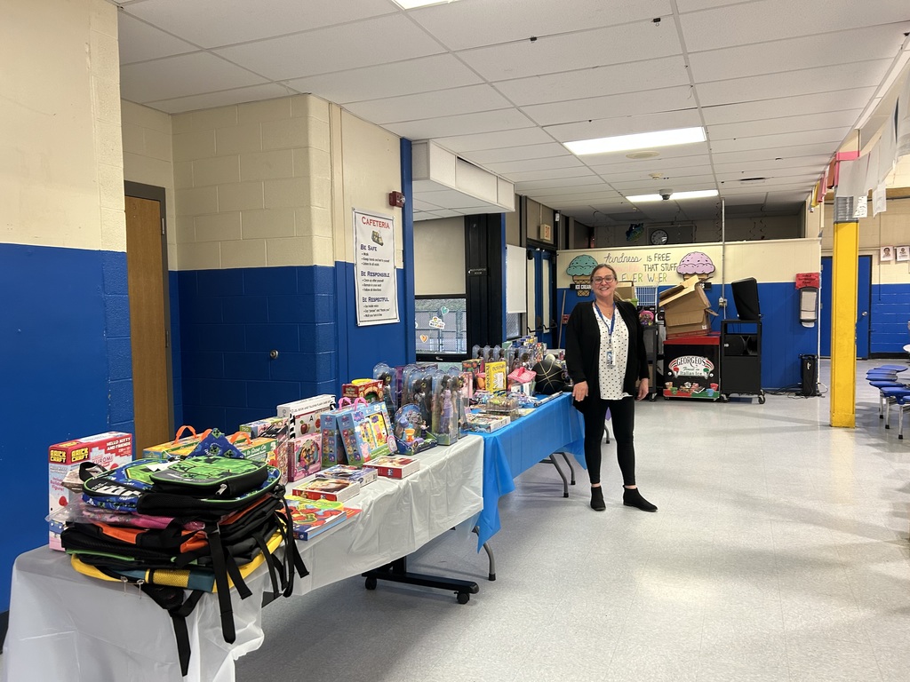 A table displays various prizes, including toys and backpacks, as a staff member stands nearby in a school cafeteria.