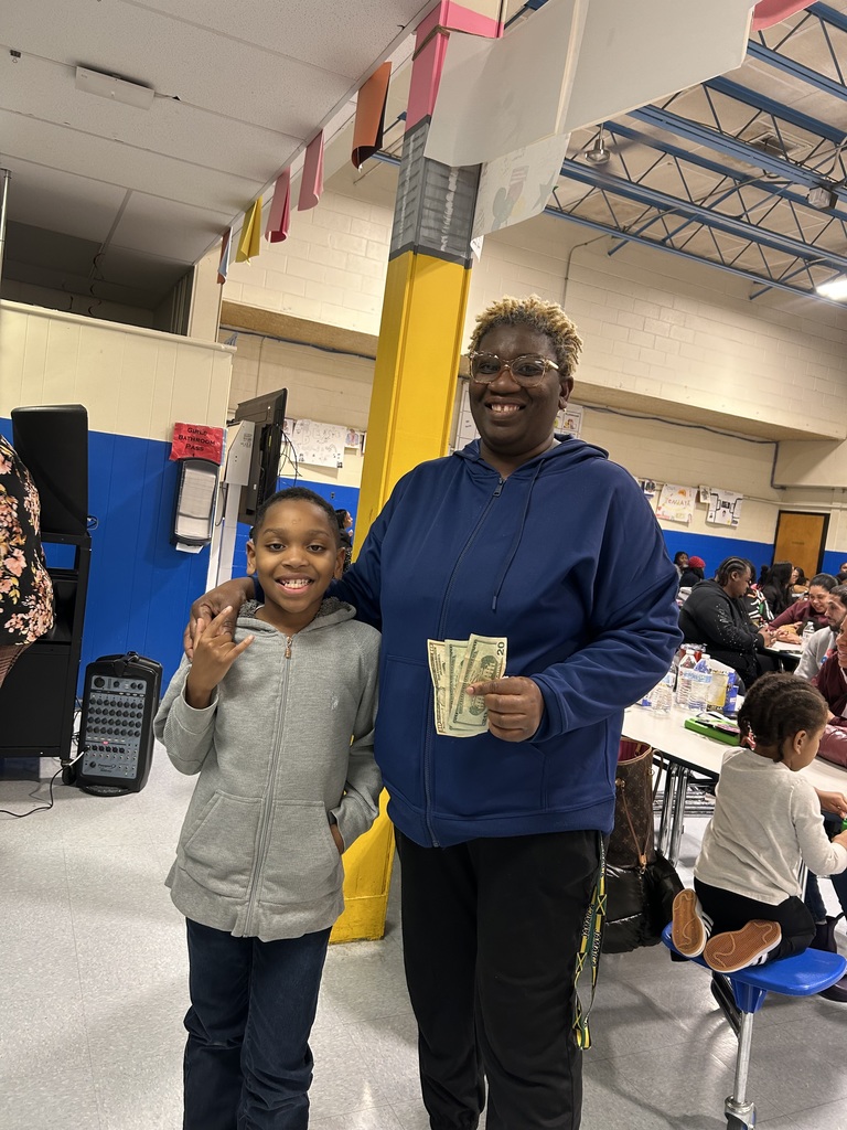 A student and an adult smile together, holding cash in a busy school cafeteria with families seated behind them.