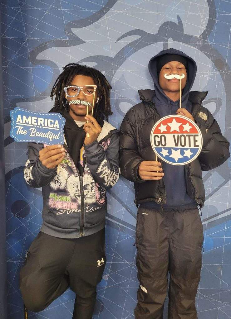 Two young men stand in front of a blue backdrop holding signs that say 'America The Beautiful' and 'Go Vote' promoting civic engagement.