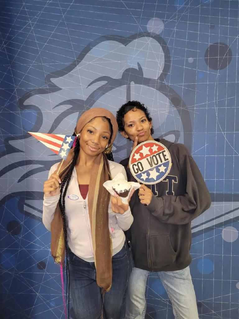 Two people posing in front of a blue wall with a knight mascot, holding a 'Go Vote' sign and an American flag cone hat.