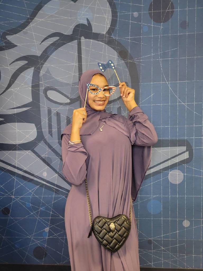 Woman in a lavender abaya and hijab poses holding star-shaped glasses against a blue geometric mural backdrop.