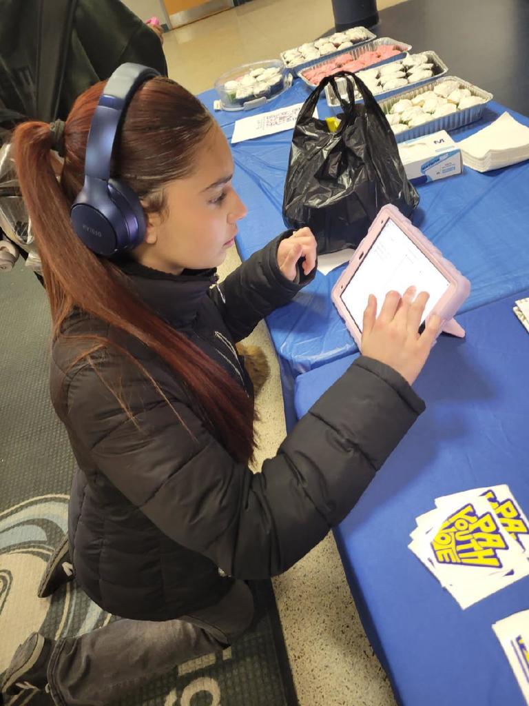 Person wearing headphones and a black jacket interacts with a tablet at a table covered with blue cloth and various items.