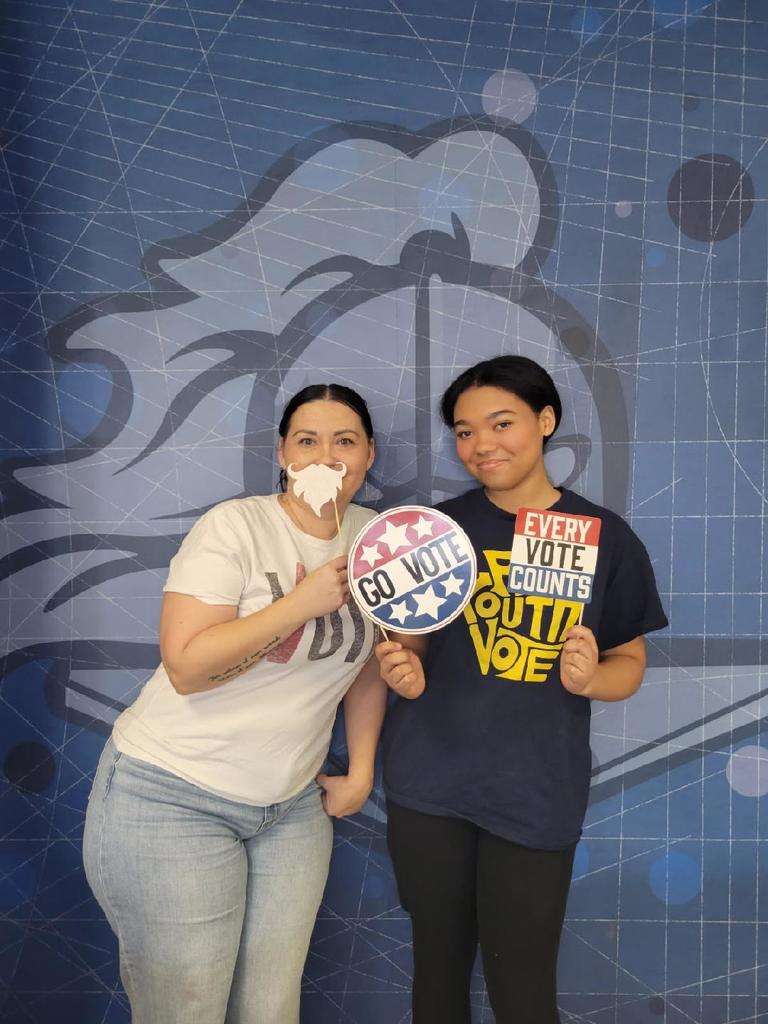 Two people standing in front of a blue backdrop holding signs that say 'GO VOTE' and 'EVERY VOTE COUNTS, GO OUT AND VOTE'.