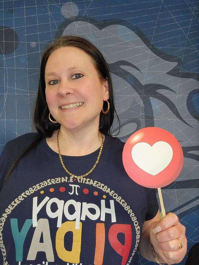 Person holding a red heart-shaped social media like sign, wearing a colorful Happy Pi Day shirt with the pi symbol and digits around the collar.