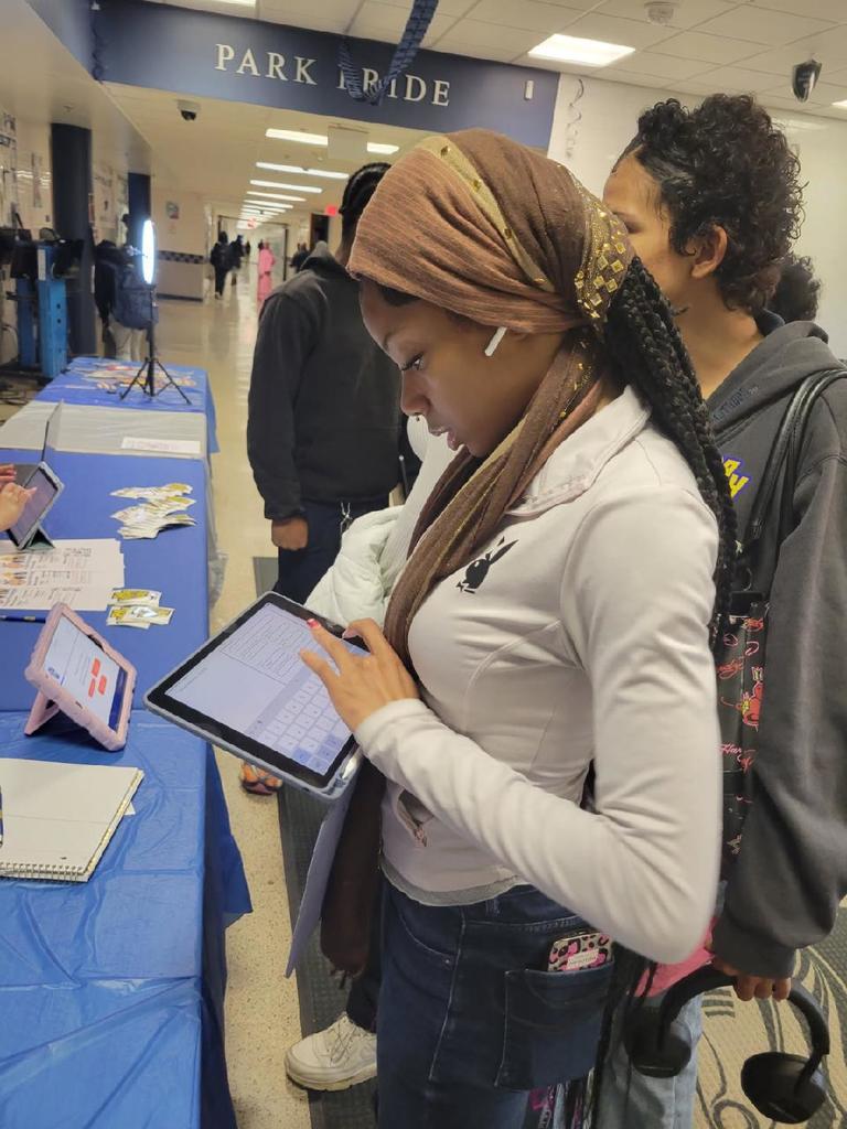 Student wearing a brown headscarf interacts with a tablet at a school event table covered in blue cloth with informational materials.