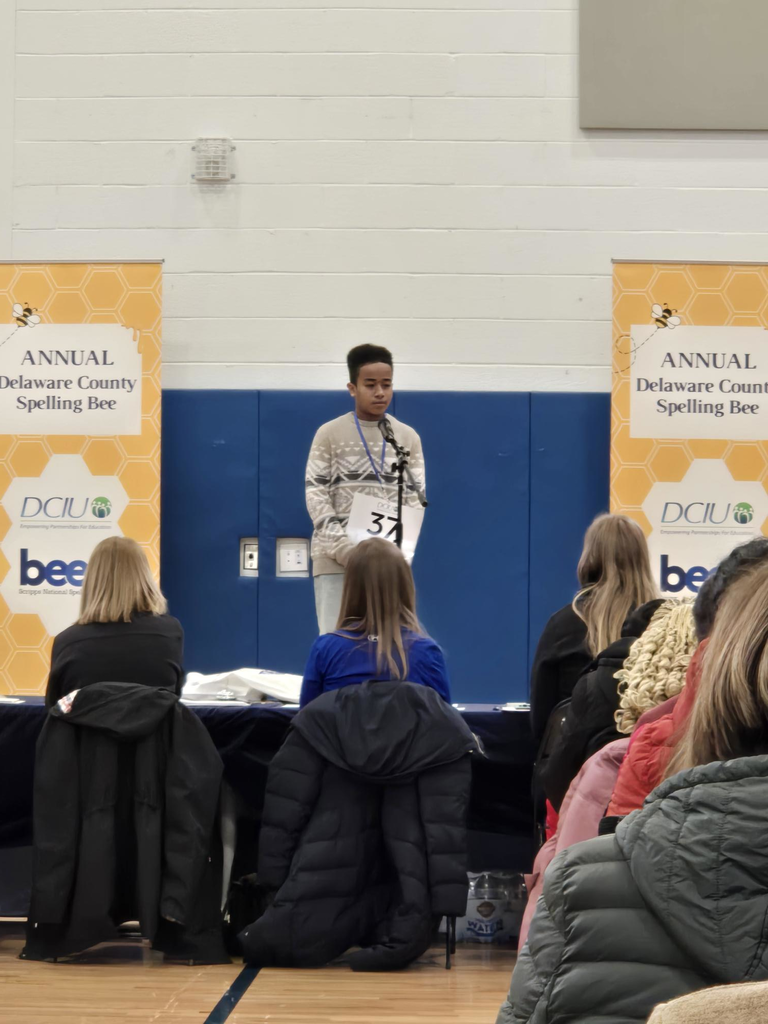 A young contestant stands at a microphone during the Annual Delaware County Spelling Bee in a gymnasium setting.