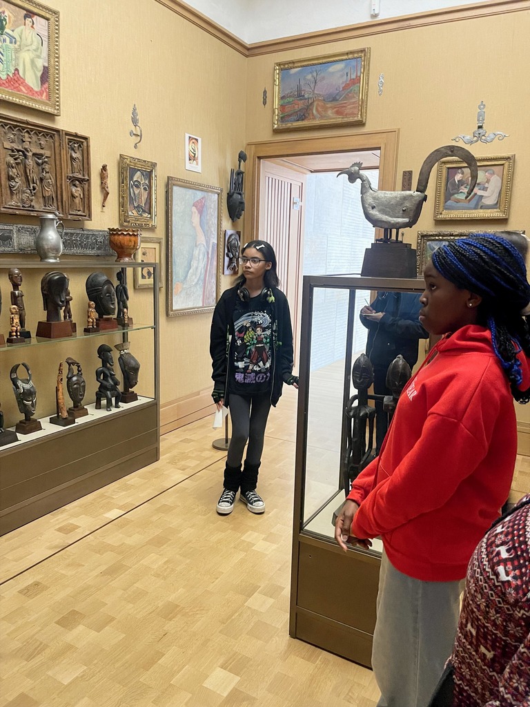 Students stand quietly in a gallery, observing carved sculptures and paintings arranged on shelves and walls.