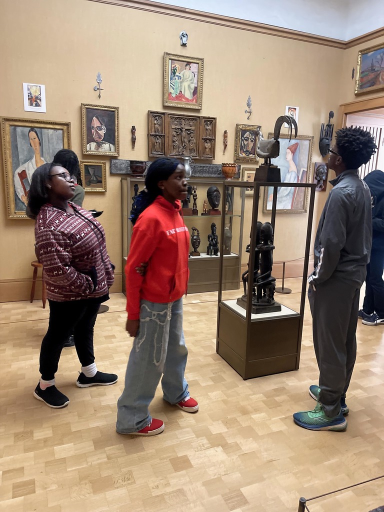 Three people attentively observing African art sculptures and framed paintings in a museum gallery with wooden flooring.
