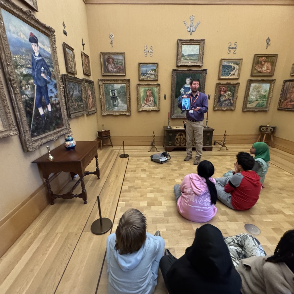 Museum educator presents to seated students, gesturing toward framed paintings while leading a discussion in an art gallery.