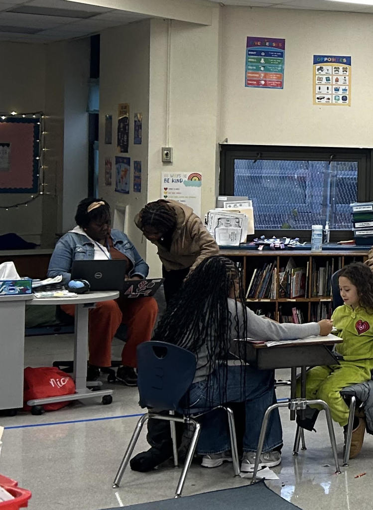Teacher assisting a student with schoolwork while another student and adult work nearby in a classroom setting.