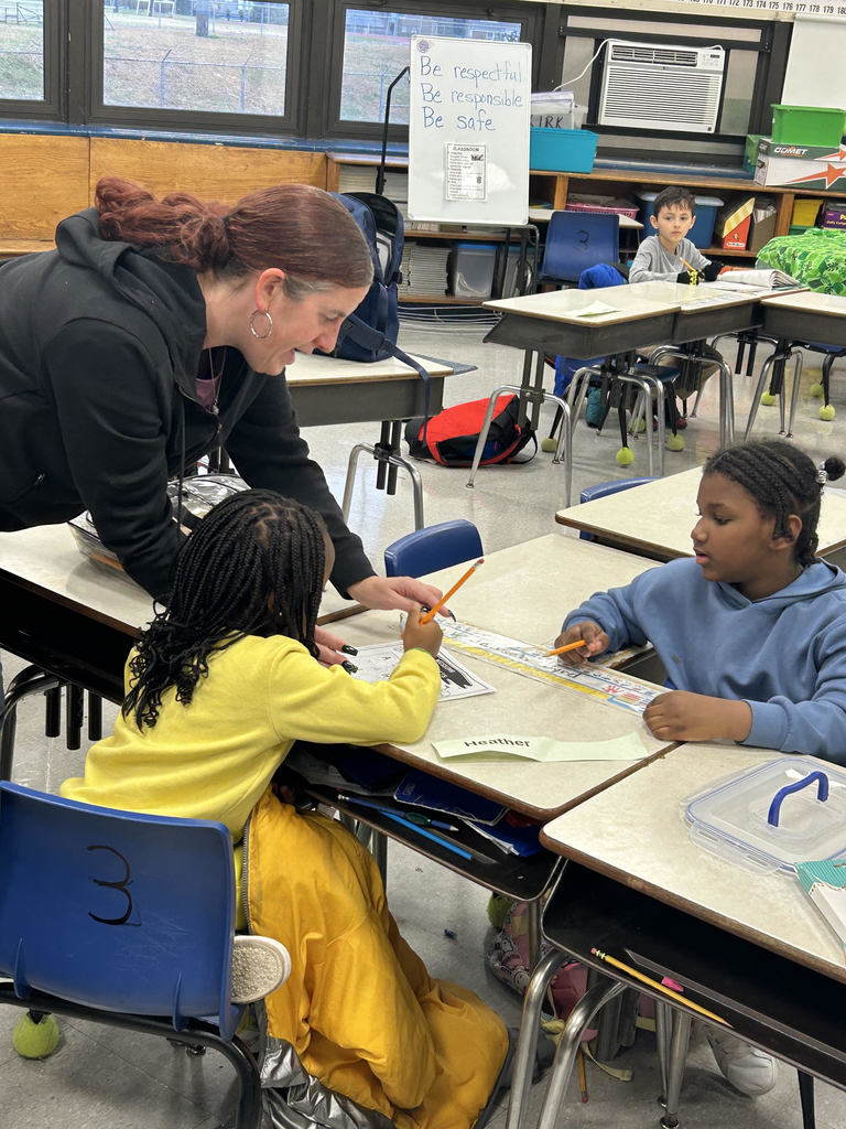 Teacher assisting young students with a classroom activity at their desks, with a whiteboard displaying classroom rules in the background.