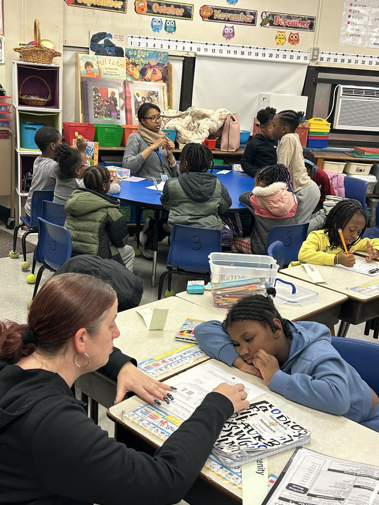 A teacher assists a young student with writing at a classroom desk while other children engage in group activities nearby.