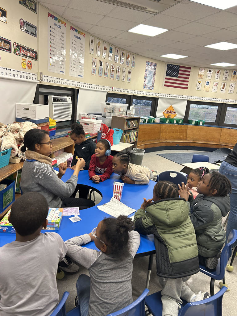 A group of young children gathered around a blue table in a classroom, engaging in a learning activity with an adult.