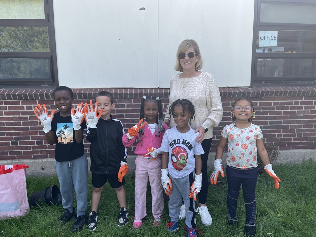 Students and staff stand outside a school holding small pots and wearing gloves, smiling during a planting or gardening activity.