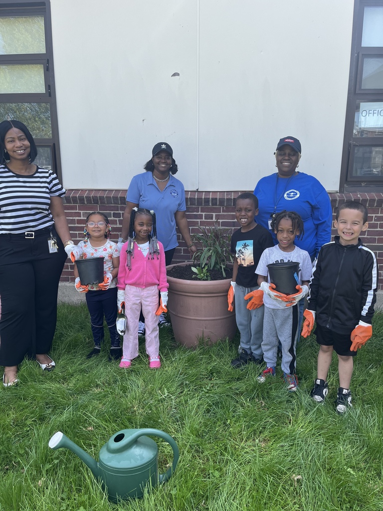 Students and staff stand outside a school holding small pots and wearing gloves, smiling during a planting or gardening activity.