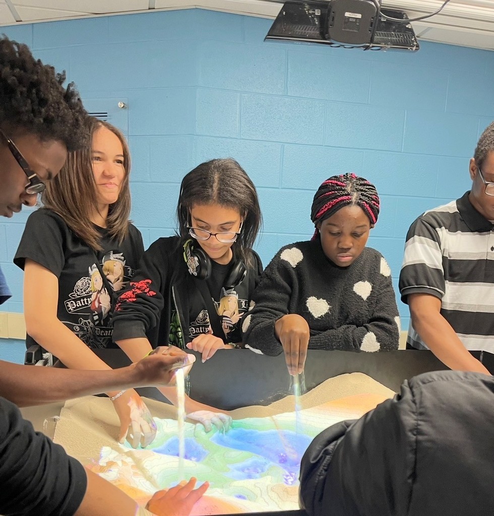 Group of children interacting with a colorful sand table, using tools to shape and explore the illuminated surface indoors.