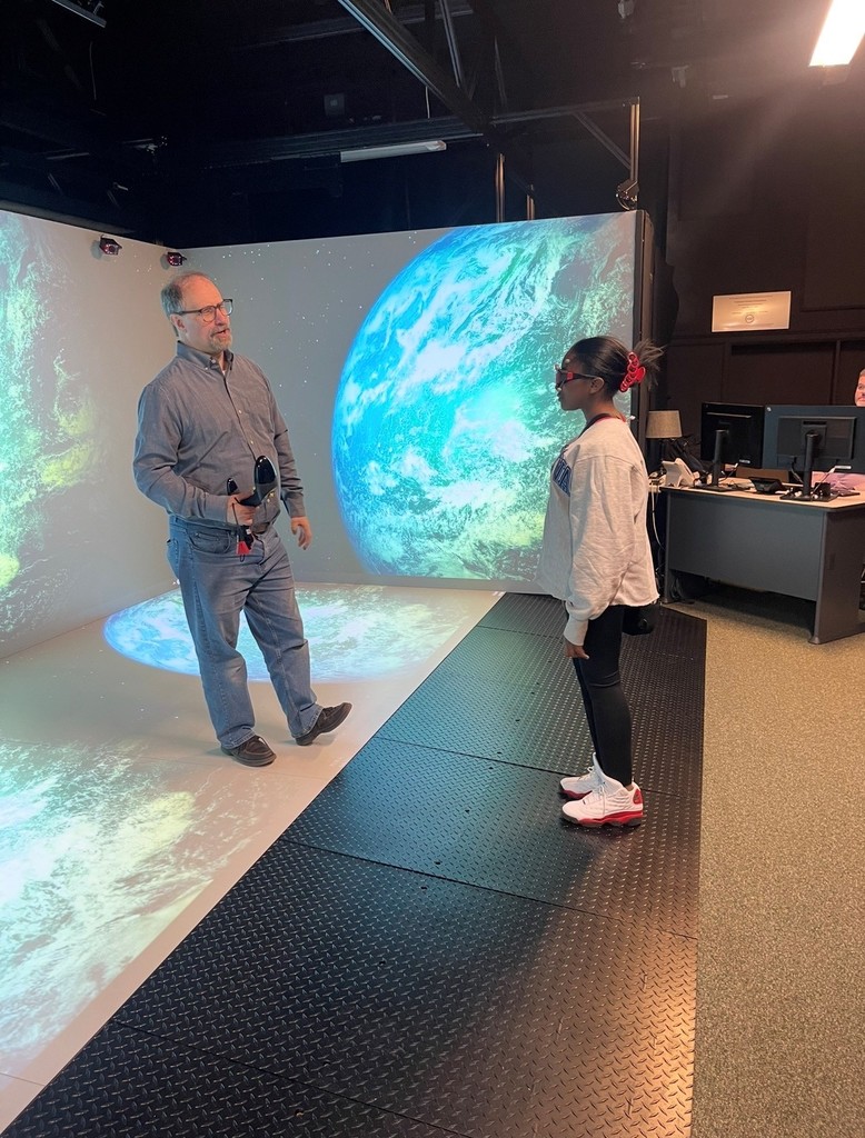 Man holding a microphone stands on a floor projection of Earth while a girl in white sneakers watches attentively in a dark room.
