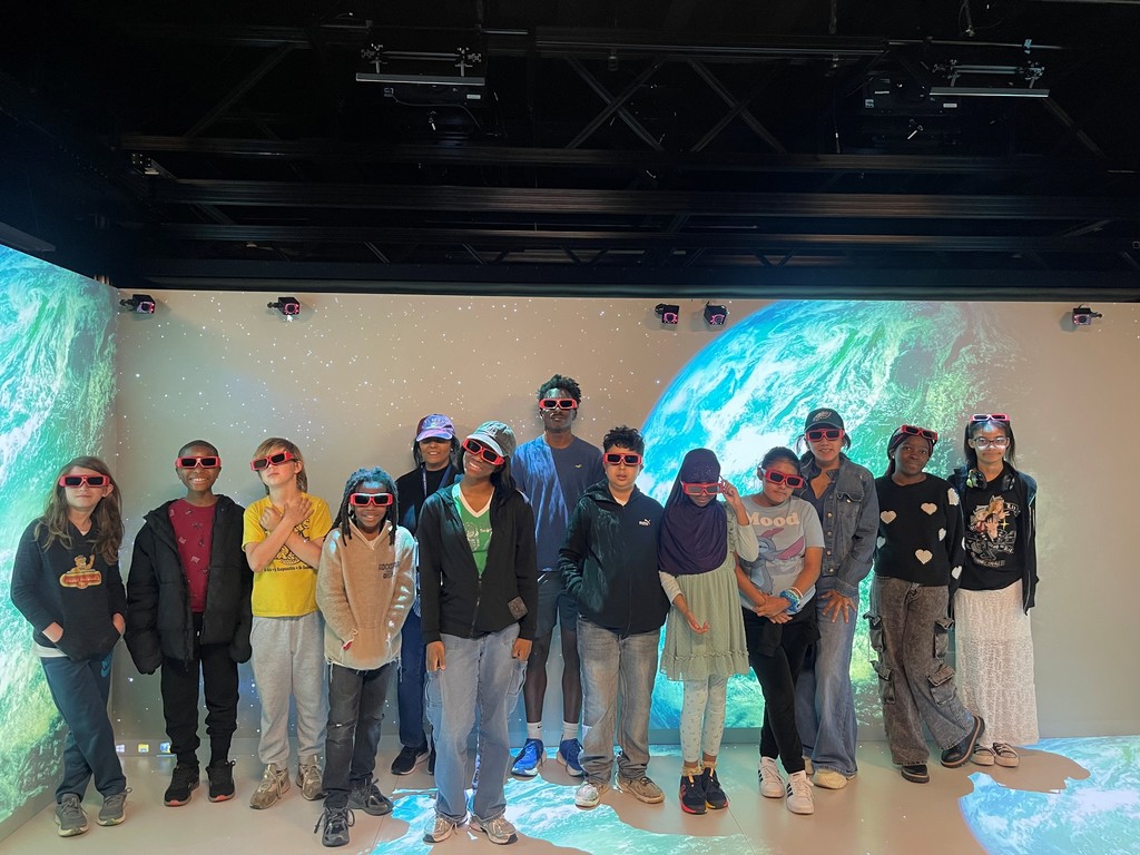 Group of children and adults wearing 3D glasses standing in front of a large Earth projection in a dark room.