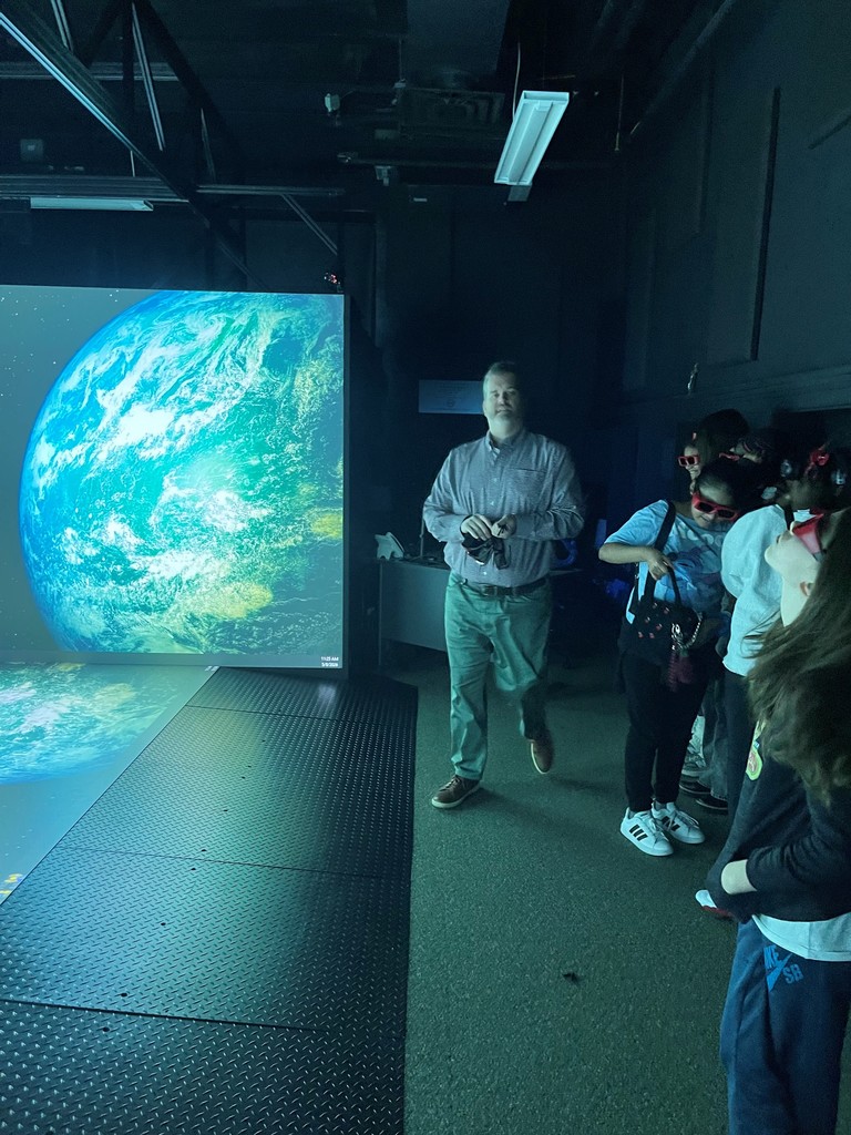 Man standing near a large illuminated globe projection in a dark room with a group of children wearing 3D glasses.