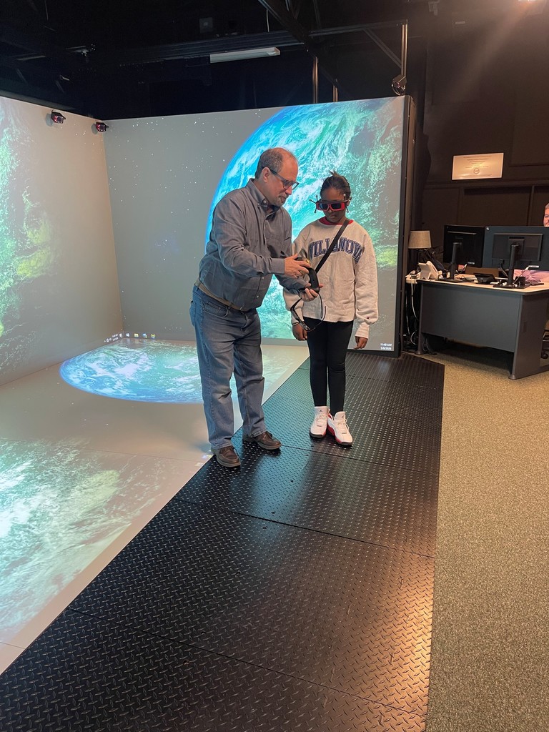 Man guiding a young girl wearing VR goggles in a room with Earth images projected on walls and floor.
