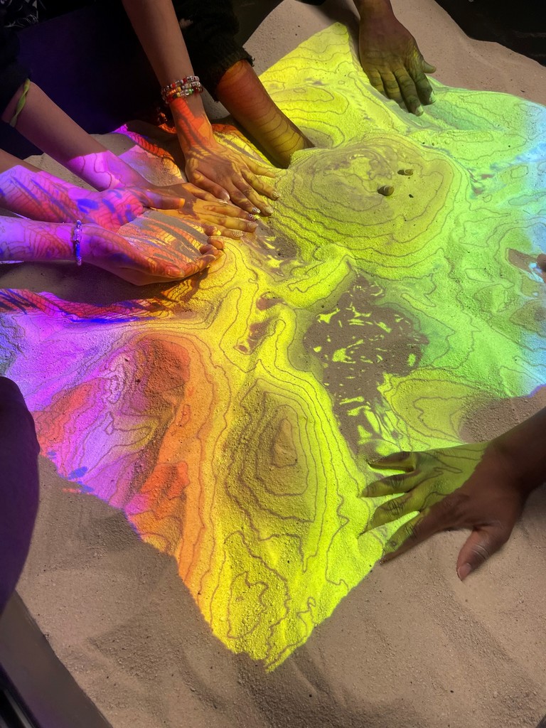 Multiple hands interact with a colorful topographic map projected onto sand, highlighting elevation contours in vibrant hues.