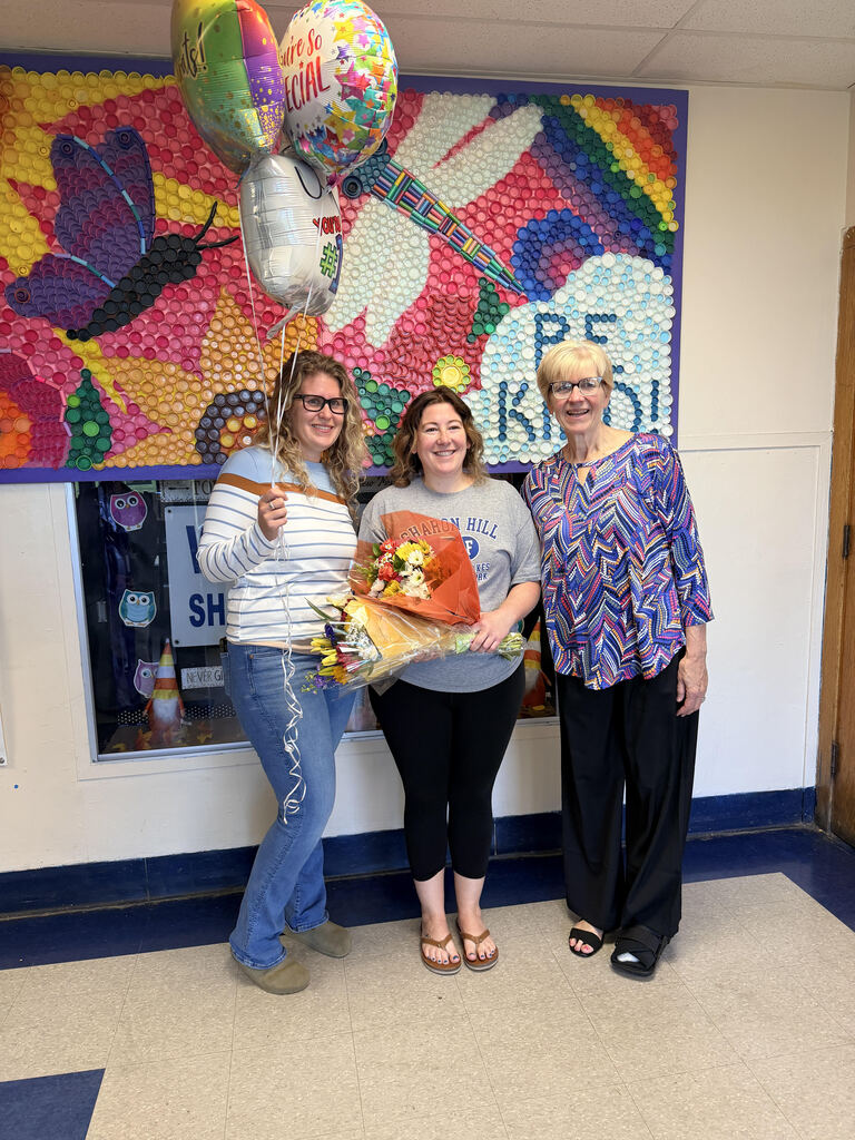Three women standing in a hallway with colorful balloon art behind them, one holding a bouquet and balloons celebrating a special occasion.