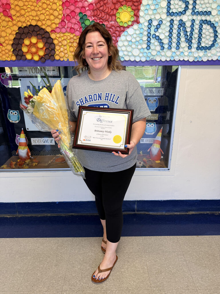 Person holding a certificate and a bouquet of yellow flowers standing in front of a colorful bulletin board with the words 'Be Kind'.