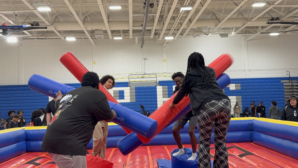 Students compete on an inflatable jousting arena, balancing on platforms while swinging padded batons.