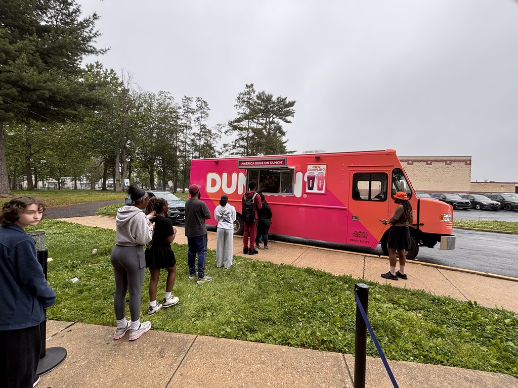 Students wait in line outside a bright pink Dunkin’ truck during a school celebration on a cloudy day.