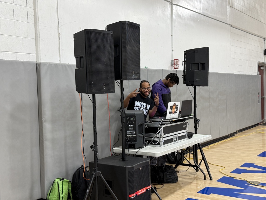 Two DJs stand behind a sound system and laptop setup in a gymnasium, providing music for an event.