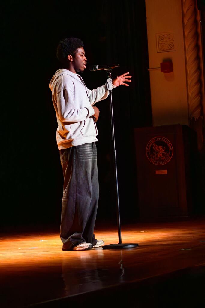 Person wearing a hoodie and jeans speaking passionately into a microphone on a dimly lit stage.